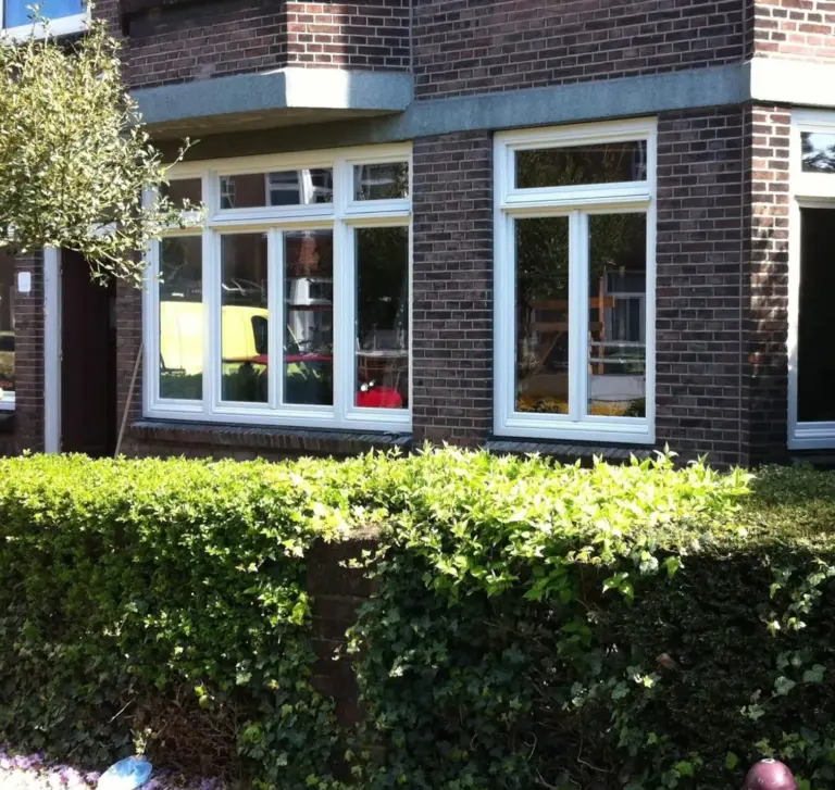 Een traditioneel Nederlands bakstenen huis met witte raamkozijnen, omgeven door strak gesnoeide groene heggen en tuinplanten in helder daglicht.