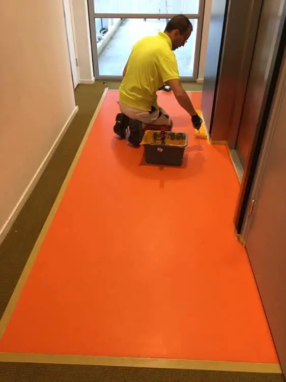Een vakman brengt met een verfroller een feloranje vloercoating aan in een binnenhal, met een gereedschapskist en schilderbenodigdheden in de buurt.