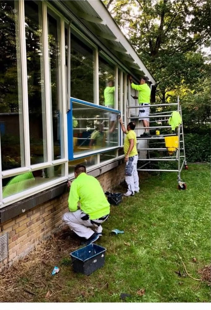 A team of four workers wearing bright neon yellow shirts and white painter's pants are installing or cleaning large windows on the side of a brick house. One worker stands on metal scaffolding near the roofline, another stands on the ground holding a window frame, and a third crouches near the foundation next to a blue bucket.