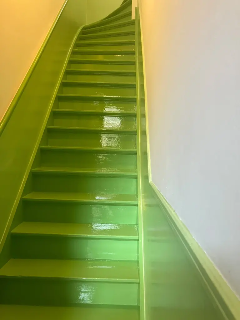 A low-angle shot looking up a winding staircase painted in a glossy, vibrant lime green. The paint reflects the overhead light on the steps and the solid side panel acting as a handrail. To the right is a plain white wall with a matching green baseboard.