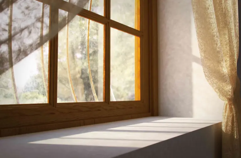Sunlight streams through a wooden window with multiple panes, casting distinct shadows across a wide, white windowsill. A sheer, patterned cream curtain hangs to the right, and blurred greenery is visible outside.