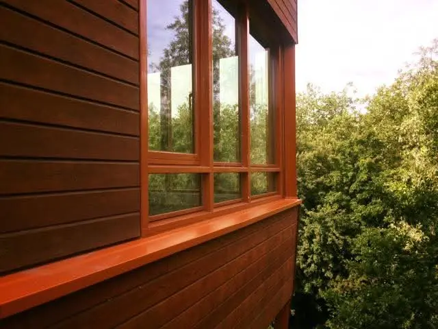 An exterior view of a building with reddish-brown horizontal wood siding and a matching wooden window frame. The glass reflects the surrounding green trees, which are also visible in the background on the right. A wide wooden windowsill runs along the bottom.